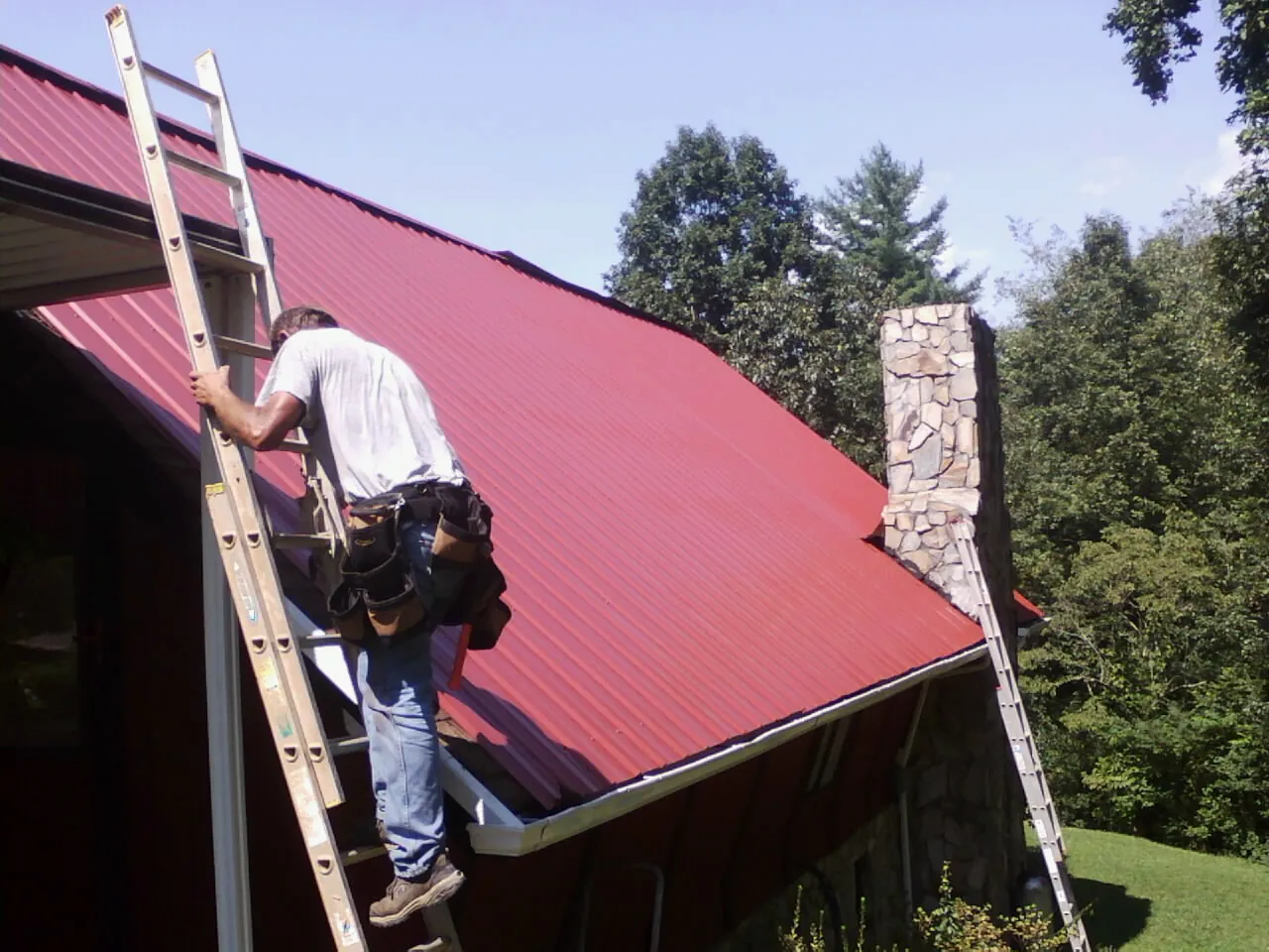 Roofer on a ladder beside a red metal roof and stone chimney — Soffit & Fascia Repair by Roofing Brothers in Sandusky